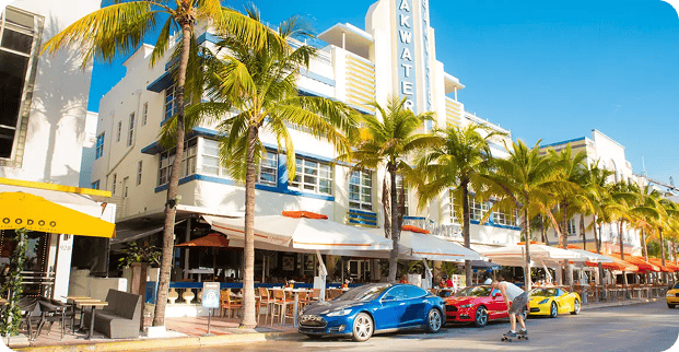 Street view of a vibrant Art Deco building on a Single Location Page, featuring outdoor dining, palm trees, parked sports cars, and a person skateboarding under a clear blue sky.